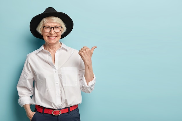 Waist up shot of friendly looking senior lady in stylish headgear, white elegant shirt and formal trousers, holds hand in pocket, points thumb away, has happy smile, advertises something nice