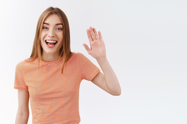 Waist-up shot friendly, pretty young woman in striped t-shirt, raising hand and waving as greeting you, lovely smiling pleasantly say hello, hi, standing white background welcoming customer in shop