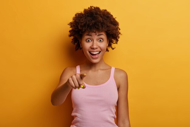 Waist up shot of funny delighted woman with Afro hairstyle points directly , sees something incredible in front, smiles positively, wears casual outfit, poses over yellow wall