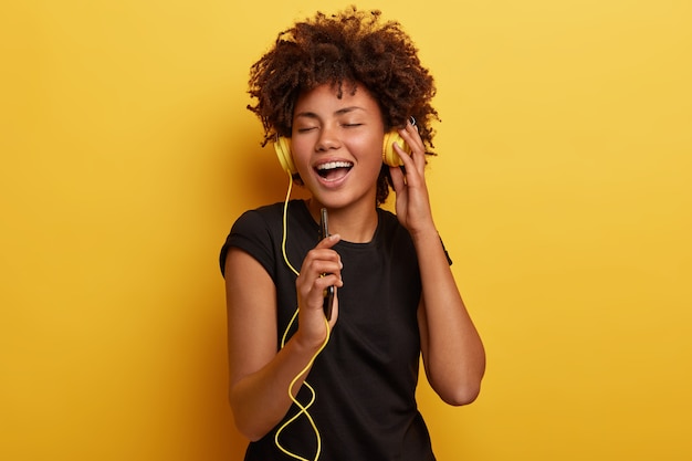 Waist up shot of glad woman with dark skin enjoys happy ending of day, listens joyful music in headphones, holds phone, has eyes shut