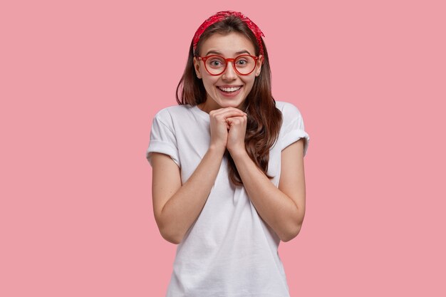 Waist up shot of glad young lady with broad smile, keeps hands together under chin, wears red headband and casual t shirt