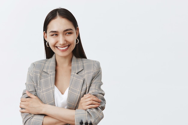 Waist-up shot of good-looking bright female in jacket and earrings, looking right and smiling broadly, holding hands crossed on chest, chuckling, having fun in team circle during business meeting