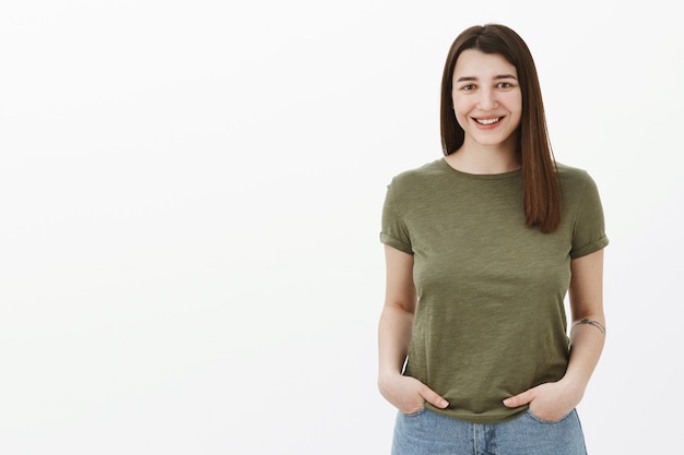 Waist-up shot of good-looking bright and sincere brunette female with short hairstyle and friendly smile grinning holding hands in pockets in casual relaxed pose over gray wall