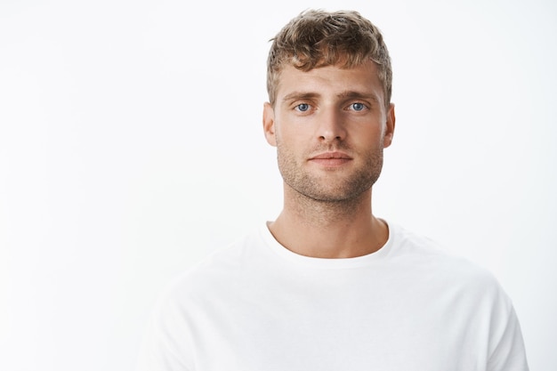 Waist-up shot of handsome calm blond blue-eyed guy with bristle in white t-shirt looking at front with relaxed carefree facial expression posing over gray wall, looking sincere and chill