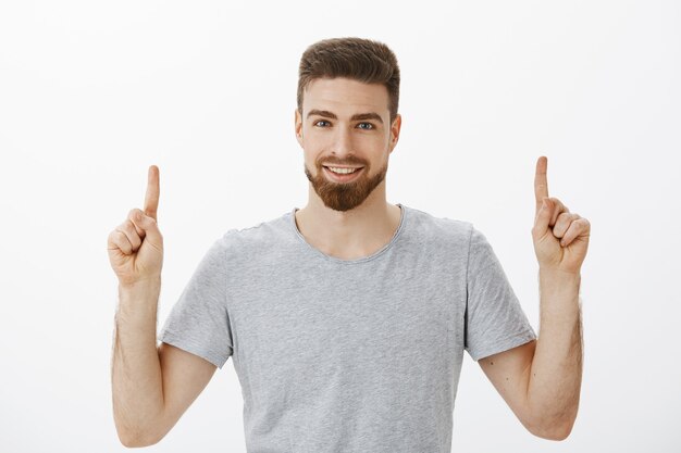 Waist-up shot of handsome self-assured male entrepreneur with beard and brown hairstyle raising index fingers pointing up with confident delighted look assuring his product is great over white wall