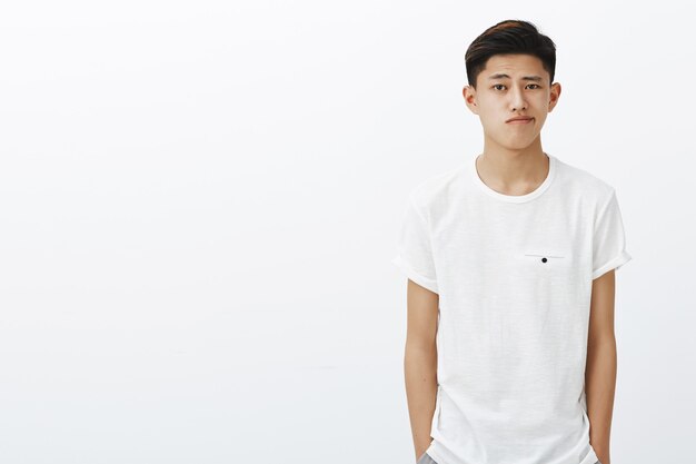 Снимок талии красивого стильного корейского парня в белой футболке, держащего руки в карманах, ухмыляющегося, стоящего в нормальной позе, тратя время зря