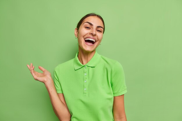 カジュアルなTシャツの笑顔に身を包んだ幸せなブルネットの女性のウエストアップショットは、緑の背景に分離されたカジュアルなTシャツを着ている面白い逸話で腕を上げて笑い続けます。幸福の概念