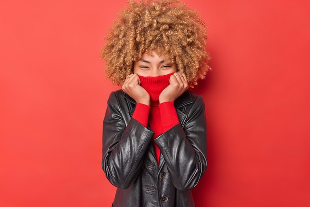 Waist up shot of happy curly haired young woman covers mouth with collar of turtleneck wears leather jacket expresses positive emotions stands against red background hides emotions foolishes around Free Photo