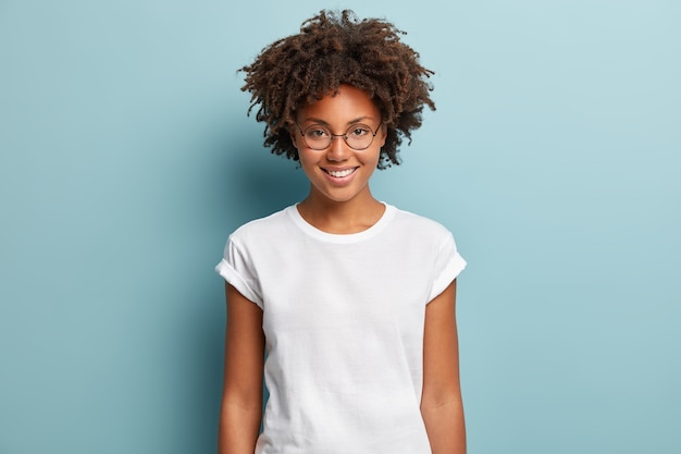 Waist up shot of happy curly woman with toothy smile, wears optical glasses and casual solid white t shirt, expresses good emotions, enjoys nice day, isolated over blue background. Face expressions
