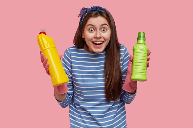 Счастливая европейская женщина с веселым выражением лица, одетая в полосатую одежду, несет бутылки с моющими средствами, подняв талию