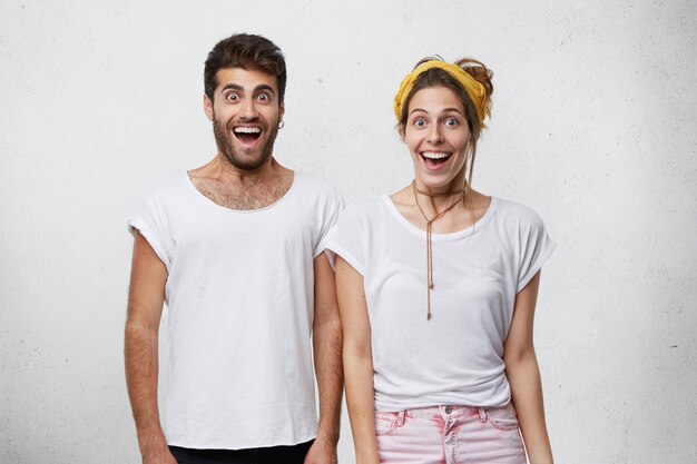 Waist up shot of happy excited man and woman dressed in white t-shirts looking in astonishment and excitement with mouths opened, rejoicing at success, victory, achievement or good news