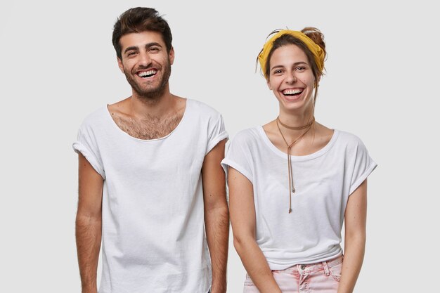 Waist up shot of happy female and male fellows dressed in white mockup t shirt, smile broadly, being in high spirit, stand closely to each other, isolated over  wall