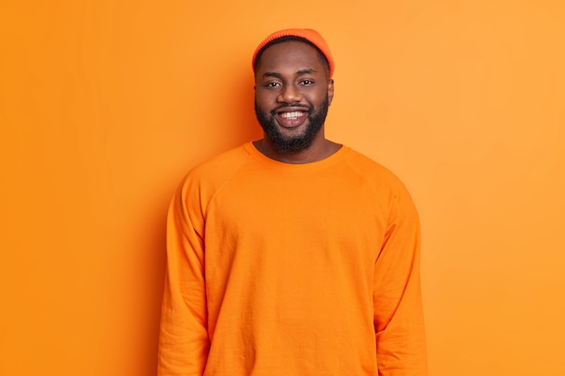 행복한 남자의 허리 위로 샷은 주황색 모자를 입고 행복하게 미소 짓고 좋은 분위기에있는 스웨터는 정면에서 직접 보이는 긍정적 인 감정을 밝은 벽에 스튜디오에 서서 표현합니다.