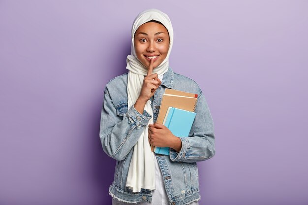 Waist up shot of happy Muslim woman with glad facial expression, makes shush gesture, shows white teeth, holds spiral notepad, wears white scarf, denim jacket, isolated over purple wall