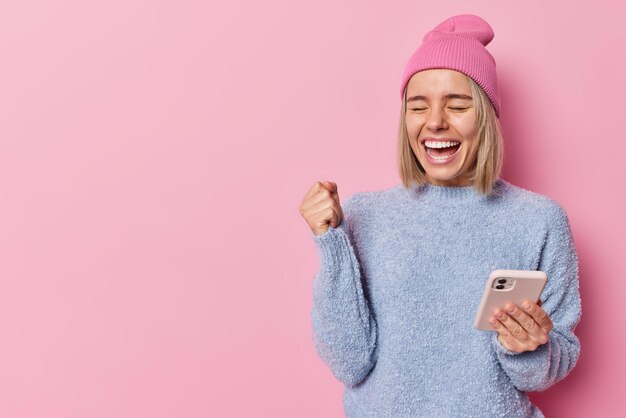 Waist up shot of happy positive blonde woman clenches fist from joy rejoices good news uses mobile phone for communication dressed in casual clothes isolated over pink background blank space away