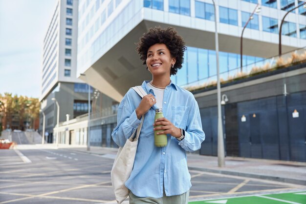 Waist up shot of happy young curly haired woman dressed in casual shirt carried fabric bag holds fresh water in bottle smiles positively strolls at city among glass skyscrapers enjoys spare time.