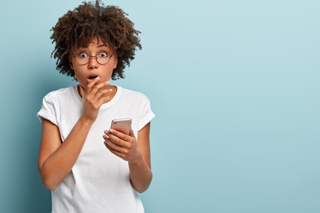 Waist up shot of impressed Afro American woman shocked to receive message with bad content, wears white t shirt, isolated over blue wall, has no internet connection, reads astonished news