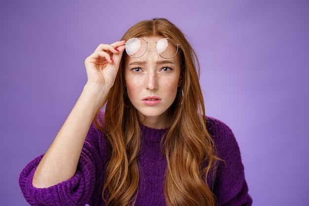 Colpo in vita di un'intensa donna rossa confusa e insicura che non può guardare senza occhiali con una brutta vista che si toglie gli occhiali e strizza gli occhi alla telecamera incerta, incapace di leggere sul muro viola.