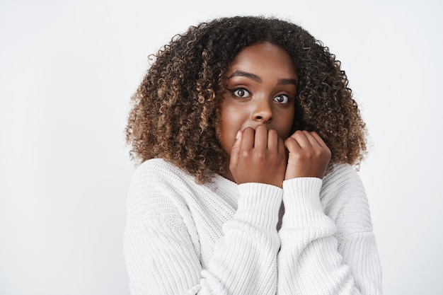 Waist-up shot of intense scared cute african-american woman watching scary movie gasping and jumping from frightening sound holding hands on lips biting fingernails nervously looking at front worried