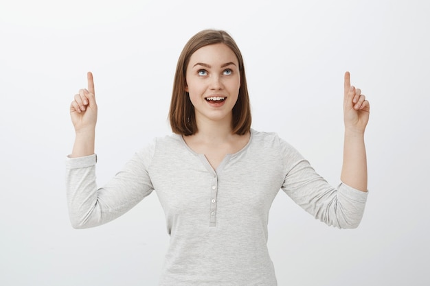 Waist-up shot of intrigued delighted good-looking european female in trendy clothes raising index fingers pointing up and smiling joyfully being interested with curious light in sky over gray wall