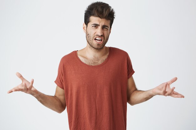 Waist up shot of irritated young man in red tshirt, trendy hairdo gesturing with hands getting angry arguing with best friend. Body language