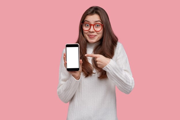Waist up shot of joyful pretty young female with dark hair points at blank screen of cell phone, shows space for your advertisement