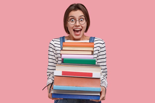 Waist up shot of joyful student in round spectacles, holds pile of books, prepares for seminar or writing report