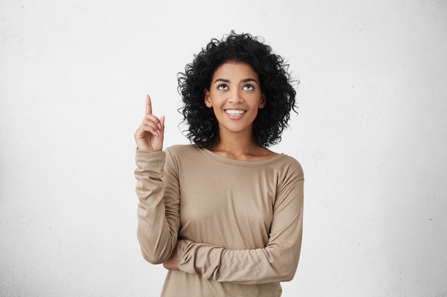 Waist up shot of joyful woman wearing beige long sleeve t-shirt looking up, pointing finger at copy space above her head. Black young woman indicating something