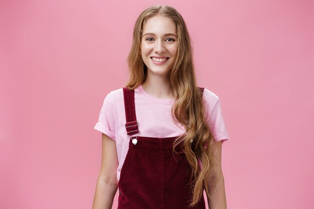 긴 물결 모양의 공정한 머리에 유행하는 작업복을 입고 즐겁게 웃고 분홍색 벽 너머로 좋은 분위기로 카메라를 응시하는 친절해 보이는 유쾌한 젊은 여학생의 웨이스트 업 샷.