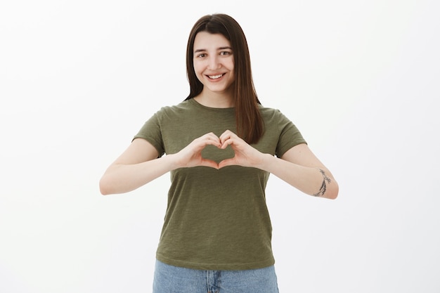 Waist-up shot of kind and sincere attractive young brunette with tattoo showing heart sign over body and smiling cute as confessing in love, making romantic gesture and sharing love