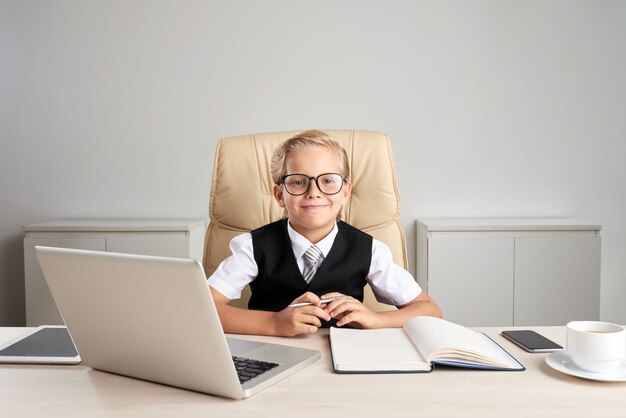 Waist up shot of little Caucasian blond boy sitting at the desk in formalwear playing the big boss