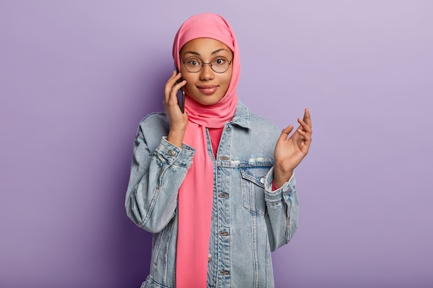 Waist up shot of Muslim female enjoys smartphone conversation, being advnaced user of modern device, wears pink hijab and denim jacket, uses public internet connection, isolated on purple wall