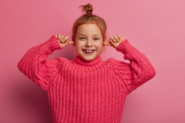 Бесплатное фото Снимок, сделанный вверх по талии, довольно радостной маленькой девочки затыкает уши указательными пальцами, радостно смеется, игнорирует громкий звук, имеет пучок волос, носит вязаный свитер, позирует на розовой стене. я тебя не слышу