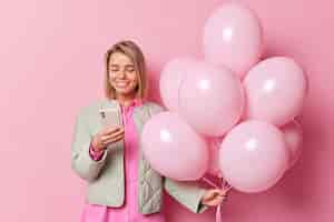 Бесплатное фото Снимок талии молодых европейских улыбающихся зубастых использует мобильный телефон, получает сообщение с поздравлениями, одетый в куртку, держит кучу надутых воздушных шаров, изолированных на розовом фоне. концепция дня рождения