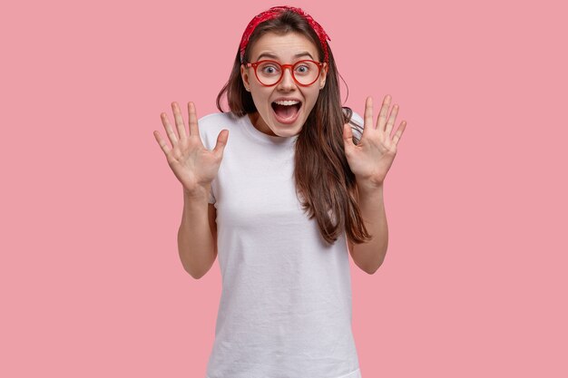 Waist up shot of overjoyed attractive woman shows palms, exclaims positively, wears spectacles and white t shirt