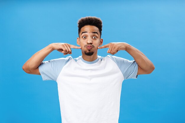 Waist-up shot of playful happy nice african american guy with tattoo on palm holding breath pouting joyfully and poking at cheeks with index fingers popping eyes while having fun over blue background