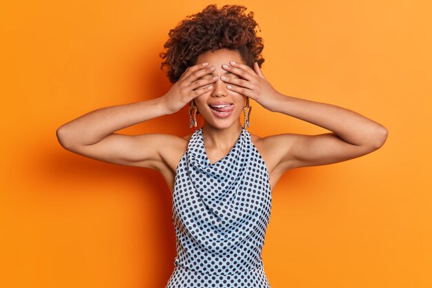 Waist up shot of playful woman covers eyes hides face sticks out tongue wears star shaped earrings and polka dot shirt isolated over orange wall