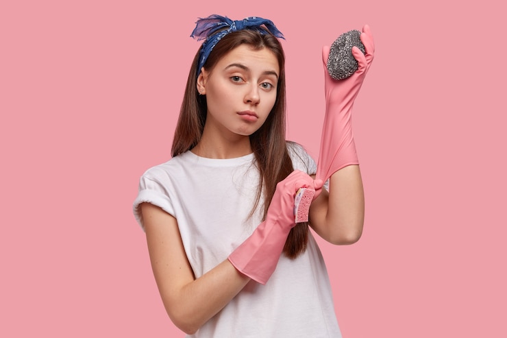 Free Photo waist up shot of pleasant looking young woman wears rubber protective gloves, uses two sponges, cleans hotel