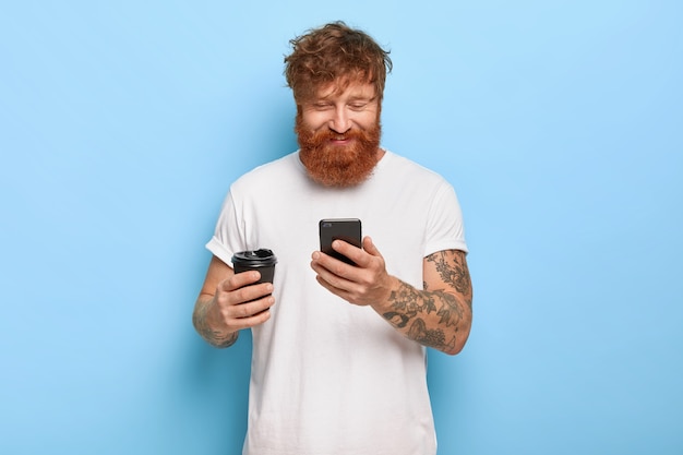 Waist up shot of pleased smiling red haired guy posing with his phone