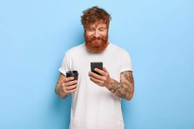 Waist up shot of pleased smiling red haired guy posing with his phone