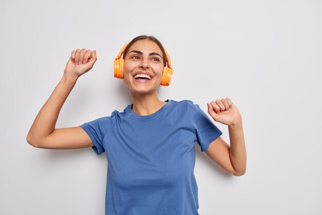 Waist up shot of positive brunette woman shakes arms catches every bit of music wears stereo headphones on ears dressed in casual blue t shirt isolated over white background enjoys favorite song