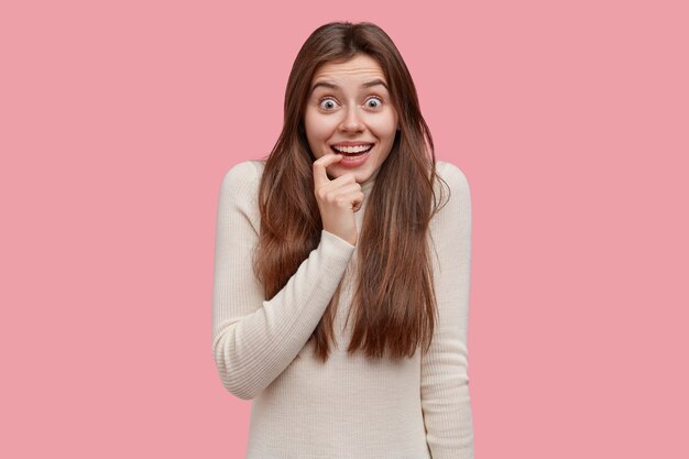 Waist up shot of positive European lady keeps finger on lips, has broad smile, looks with excitement