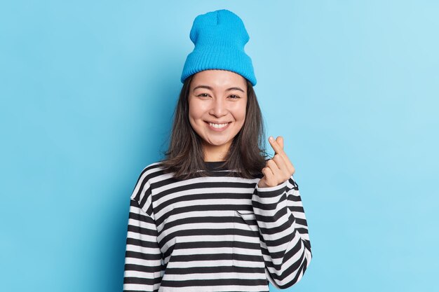 Waist up shot of pretty Asian woman with dark hair makes korean heart with fingers wears hat striped jumper smiles pleasantly 