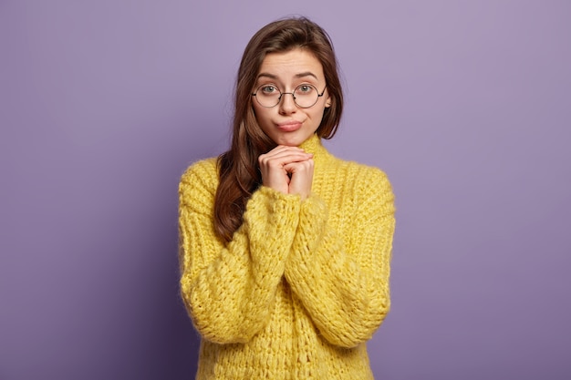 Waist up shot of pretty brunette female feels lonely and sad, keeps hands under chin, upset by bad news, wears yellow jumper, stands over purple  wall. Human facial expressions and feelings
