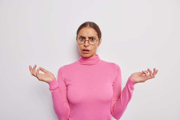 Waist up shot of pretty brunette woman spreads palms has puzzled indecisive expression reacts on something unpleasant isolated over white wall