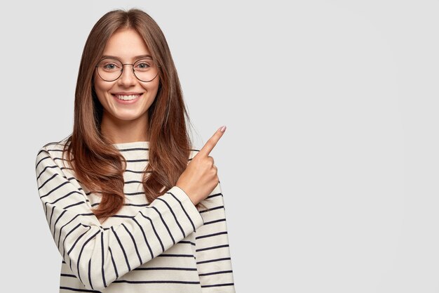 Waist up shot of pretty Caucasian woman with cheerful expression, points with index finger at blank copy space, dressed in striped sweater, shows free space at upper right corner for your promotion