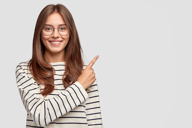 Waist up shot of pretty Caucasian woman with cheerful expression, points with index finger at blank copy space, dressed in striped sweater, shows free space at upper right corner for your promotion