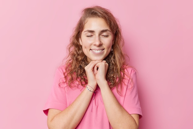 Снимок талии довольно длинноволосой молодой женщины мечтает о чем-то приятном с закрытыми глазами, держит руки под подбородком, носит повседневную футболку, изолированную на розовом фоне. Концепция счастливых чувств