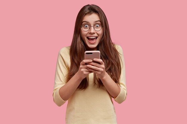 Waist up shot of pretty satisfied woman holds mobile phone, happy to exchange text messages in social networks