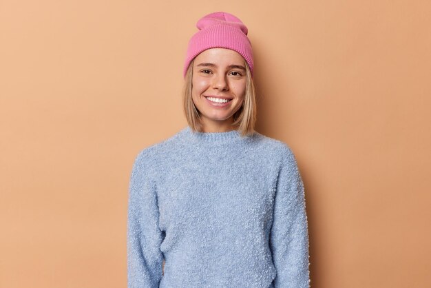 Waist up shot of pretty young woman smiles toothily looks with happy expression at camera dressed in casual jumper pink hat has good mood isolated over brown background. Positive emotions concept